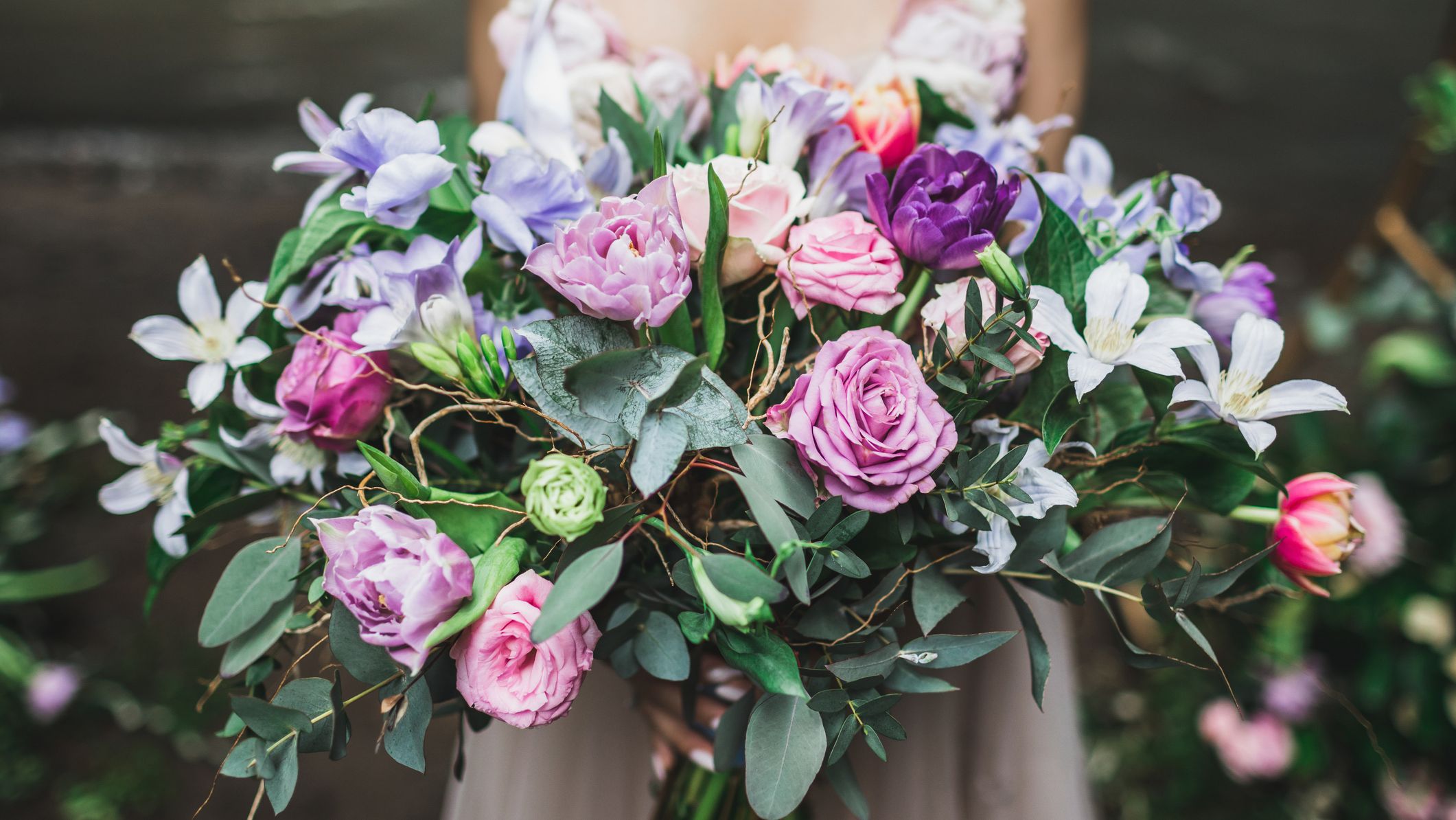 A bride holds a lush bouquet of roses, tulips, and wildflowers in shades of pink, purple, and white, accented with greenery and delicate vines.
