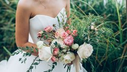 A bride in a white strapless gown holds a romantic bouquet of white and pink roses, peonies, and greenery, with delicate ribbon accents, against a lush green backdrop.