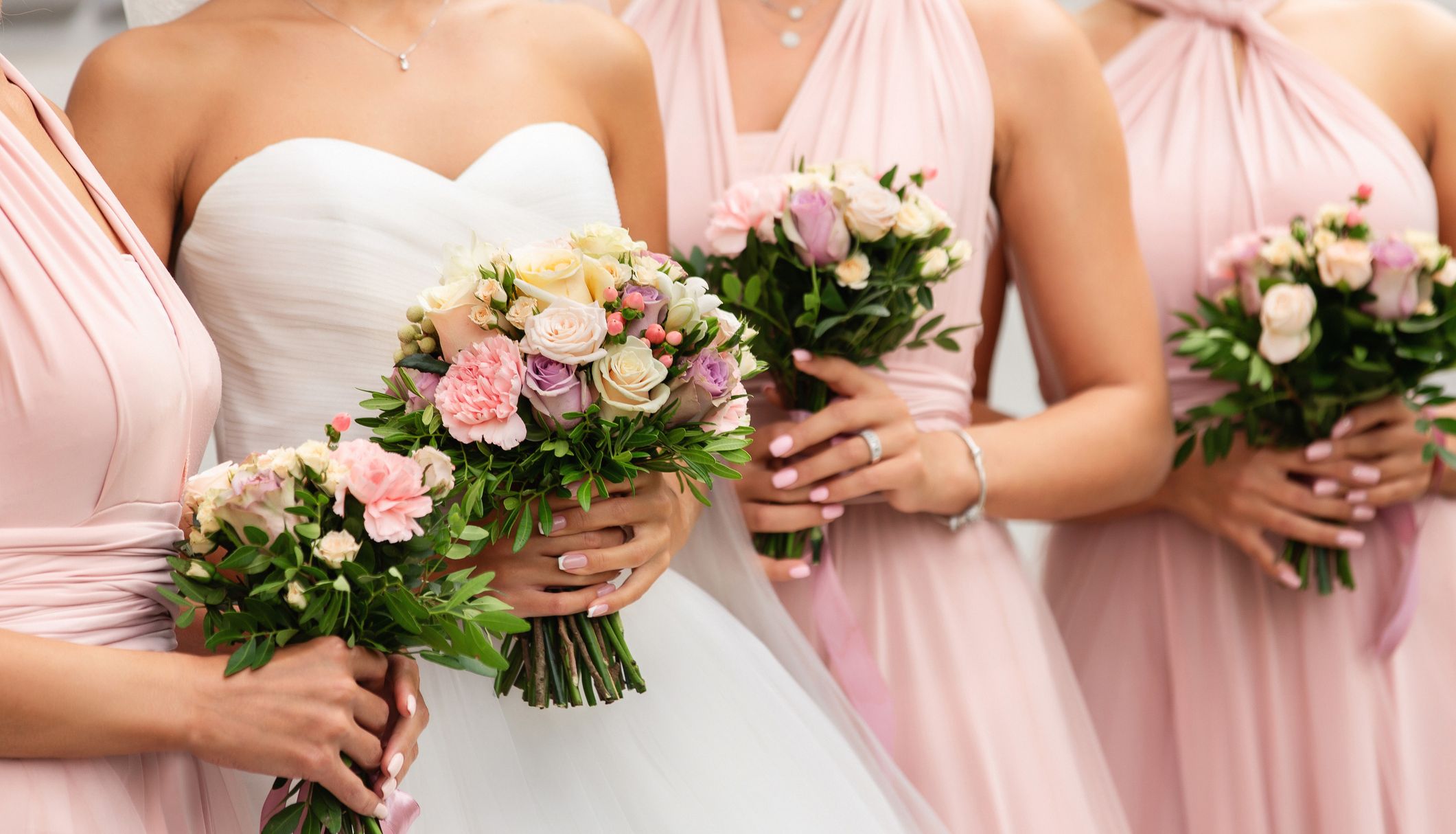 A bride in a white gown stands with her bridesmaids in blush pink dresses, all holding elegant bouquets of roses, carnations, and greenery with soft ribbon accents.
