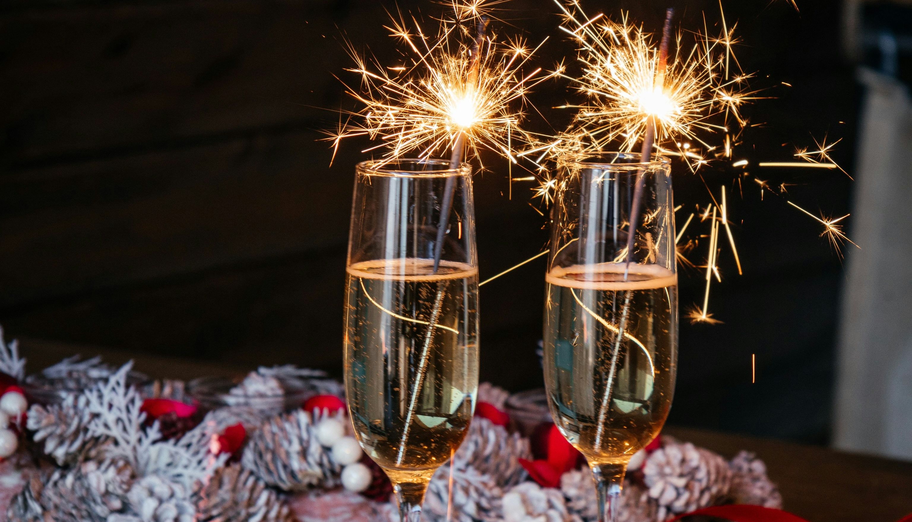 Two champagne glasses with sparklers, festive pinecone wreath and red ribbon on wooden table