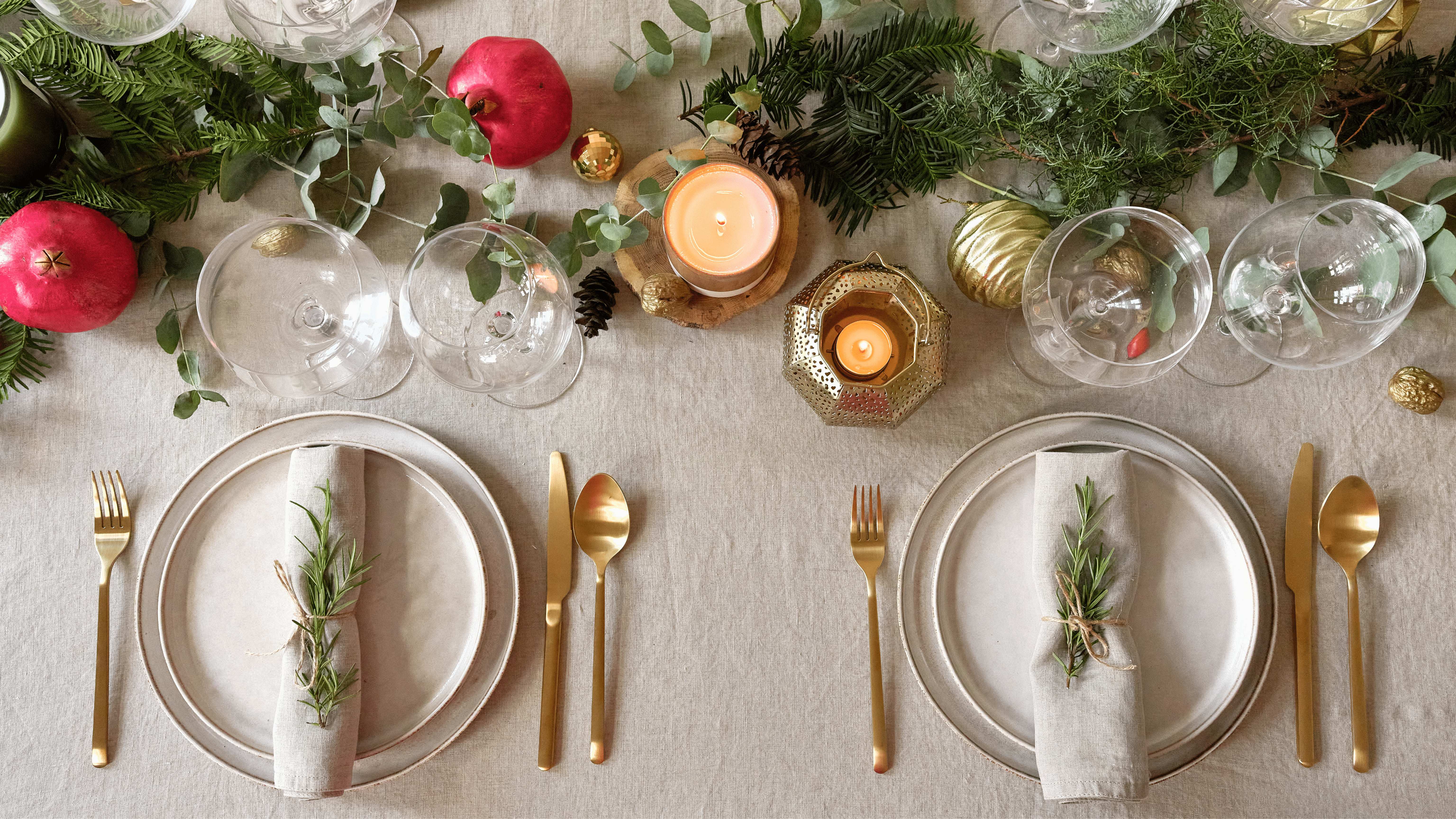 Elegant holiday table setting with gold cutlery, candles, greenery, pomegranates, and wine glasses.