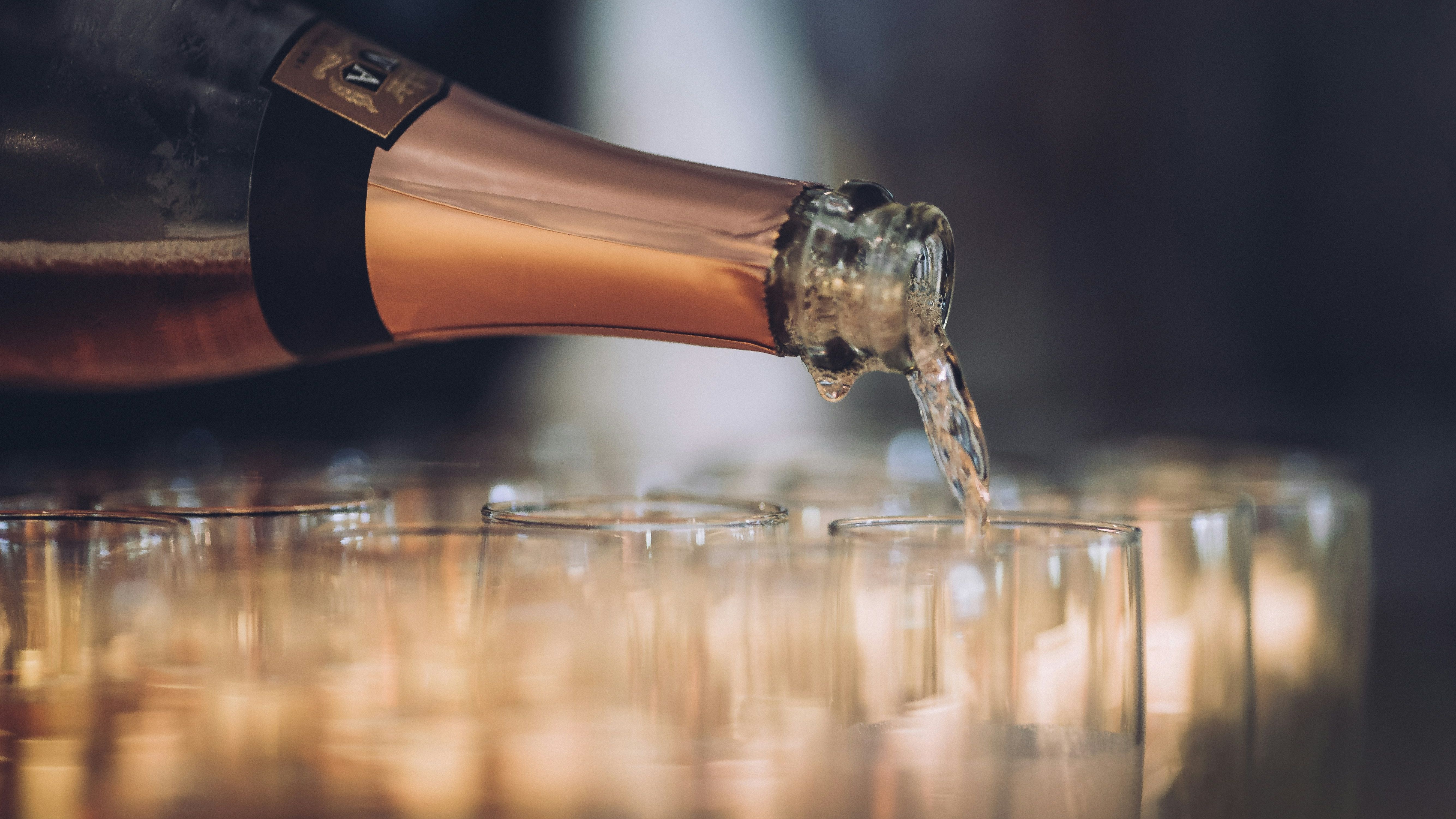 A close-up shot of a champagne bottle pouring sparkling wine into a row of clear glasses.