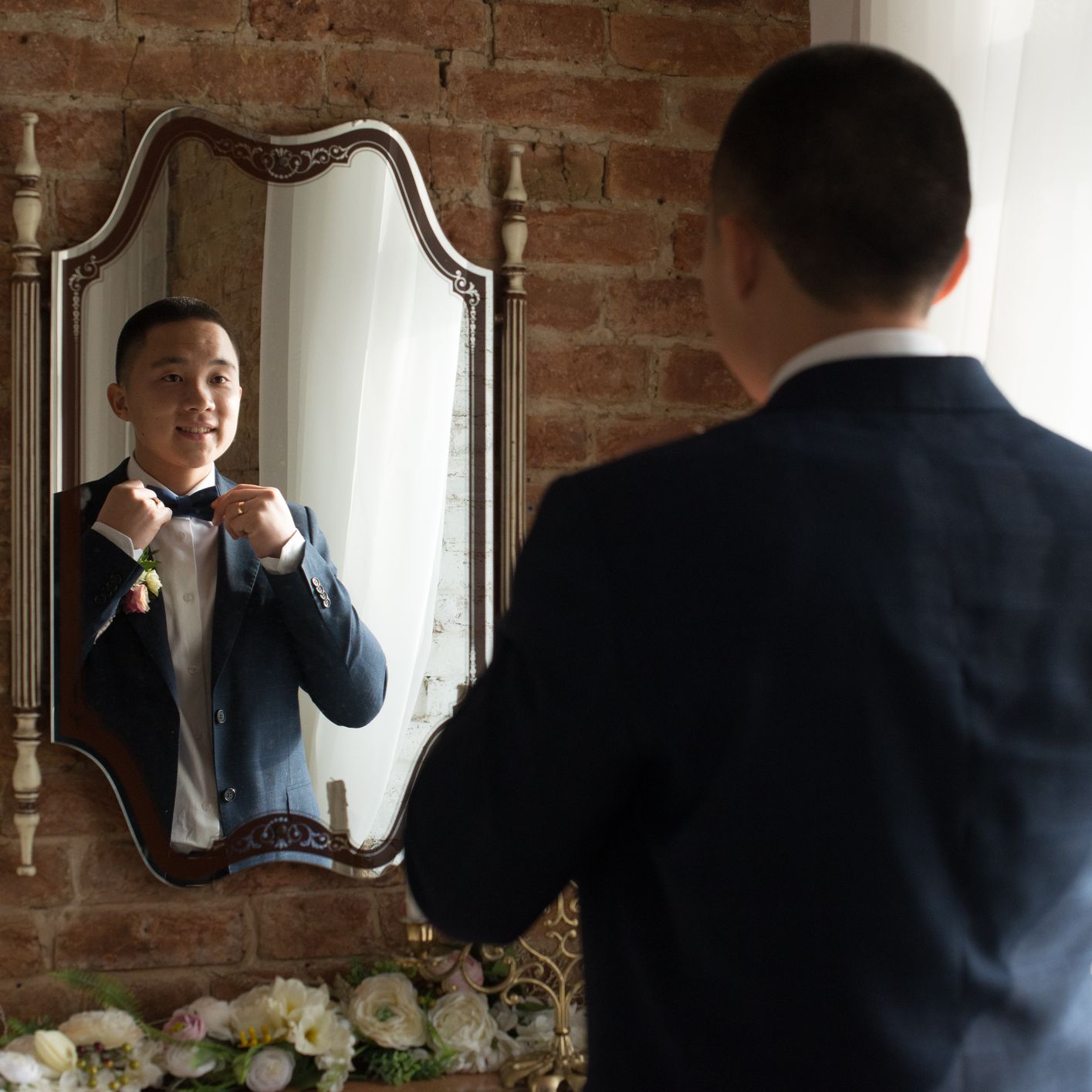 Groom adjusts his bow tie while smiling at his reflection in a mirror, standing in a softly lit room with exposed brick walls and white curtains.
