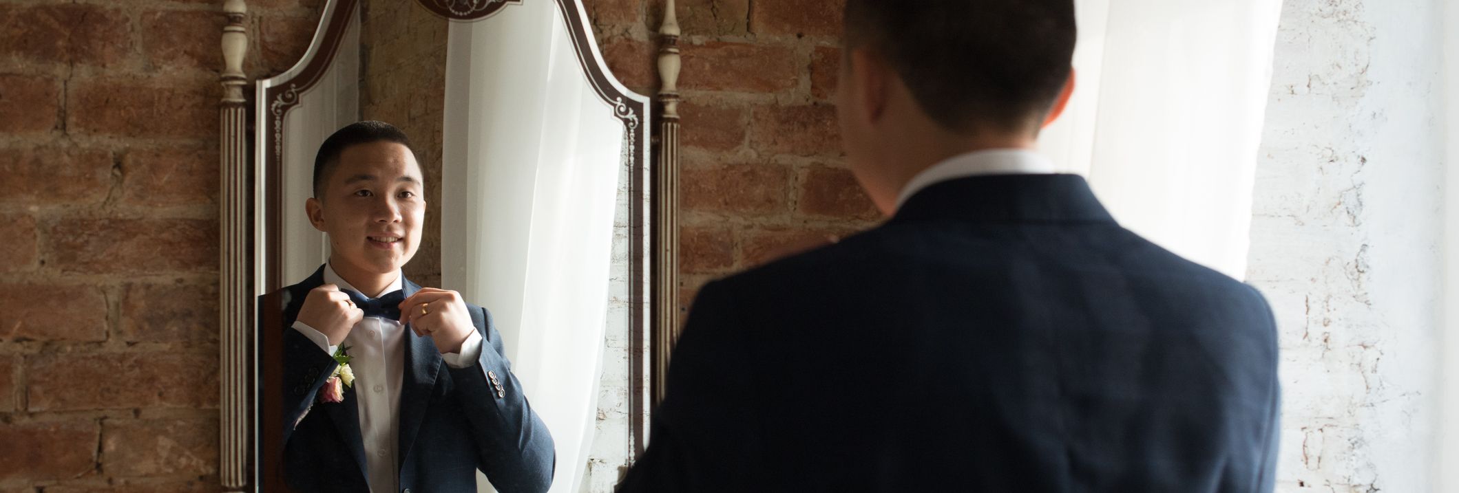Groom adjusts his bow tie while smiling at his reflection in a mirror, standing in a softly lit room with exposed brick walls and white curtains.