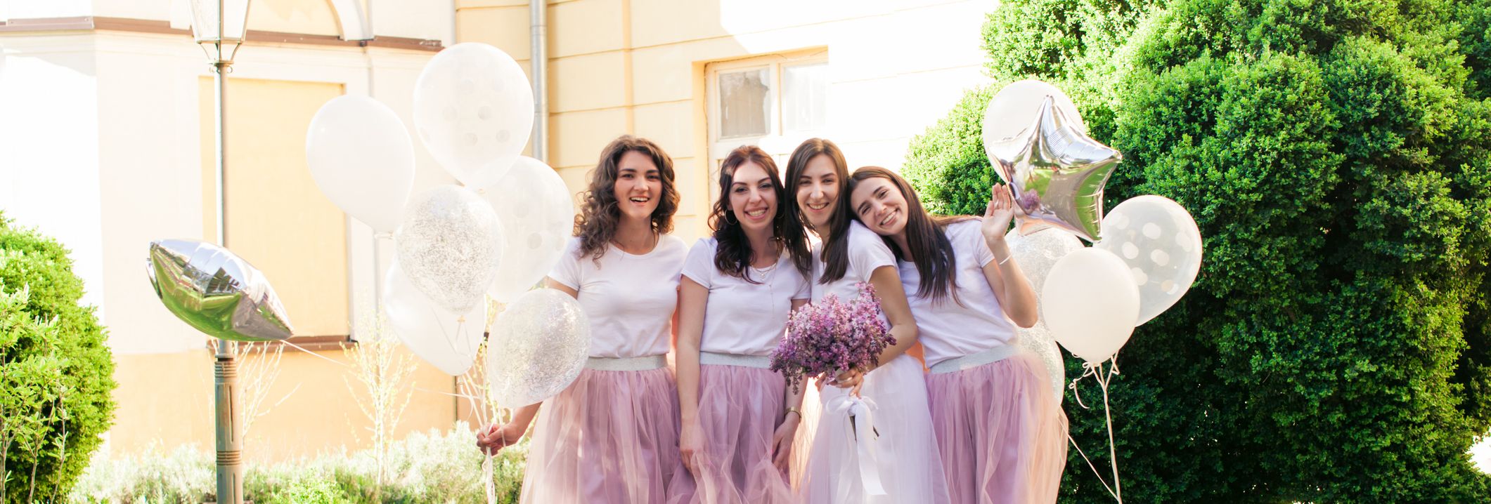 Bride and bridesmaids pose cheerfully outdoors, all wearing white tops and tulle skirts, holding balloons and flowers on a sunny cobbled path.
