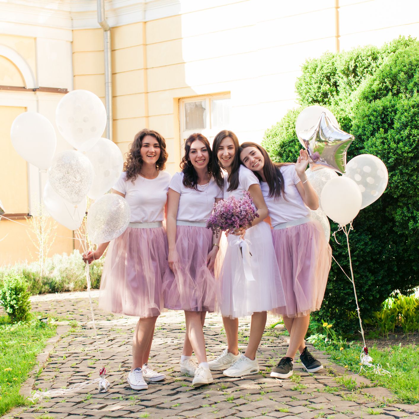 Bride and bridesmaids pose cheerfully outdoors, all wearing white tops and tulle skirts, holding balloons and flowers on a sunny cobbled path.