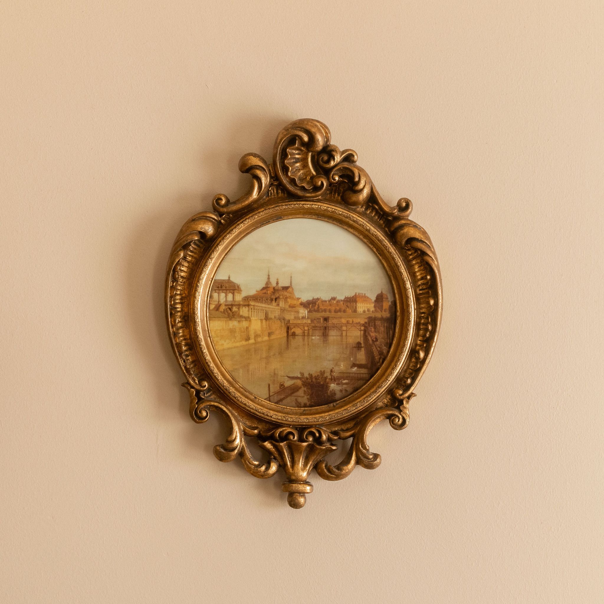 Ornate vintage gold-framed painting of a cityscape with river, hanging on a beige wall.