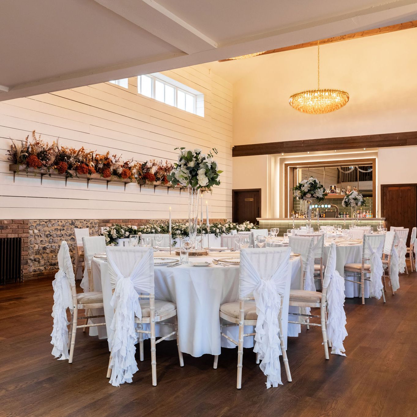 Elegant wedding reception hall with round tables decorated with white linens and ruffled chair covers, floral centerpieces, and a sparkling chandelier.