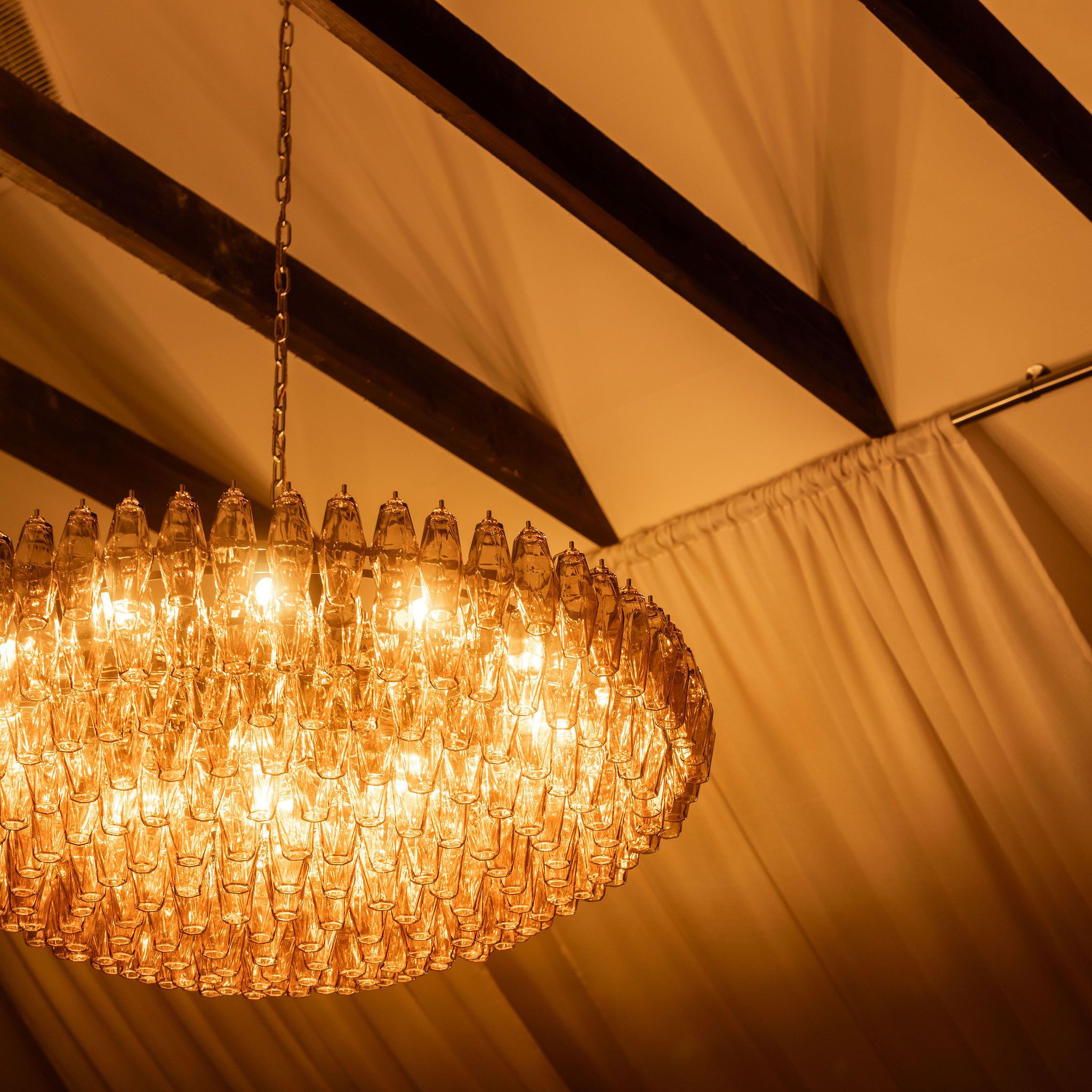Elegant chandelier with warm yellow lights hanging from a ceiling with draped fabric and exposed beams