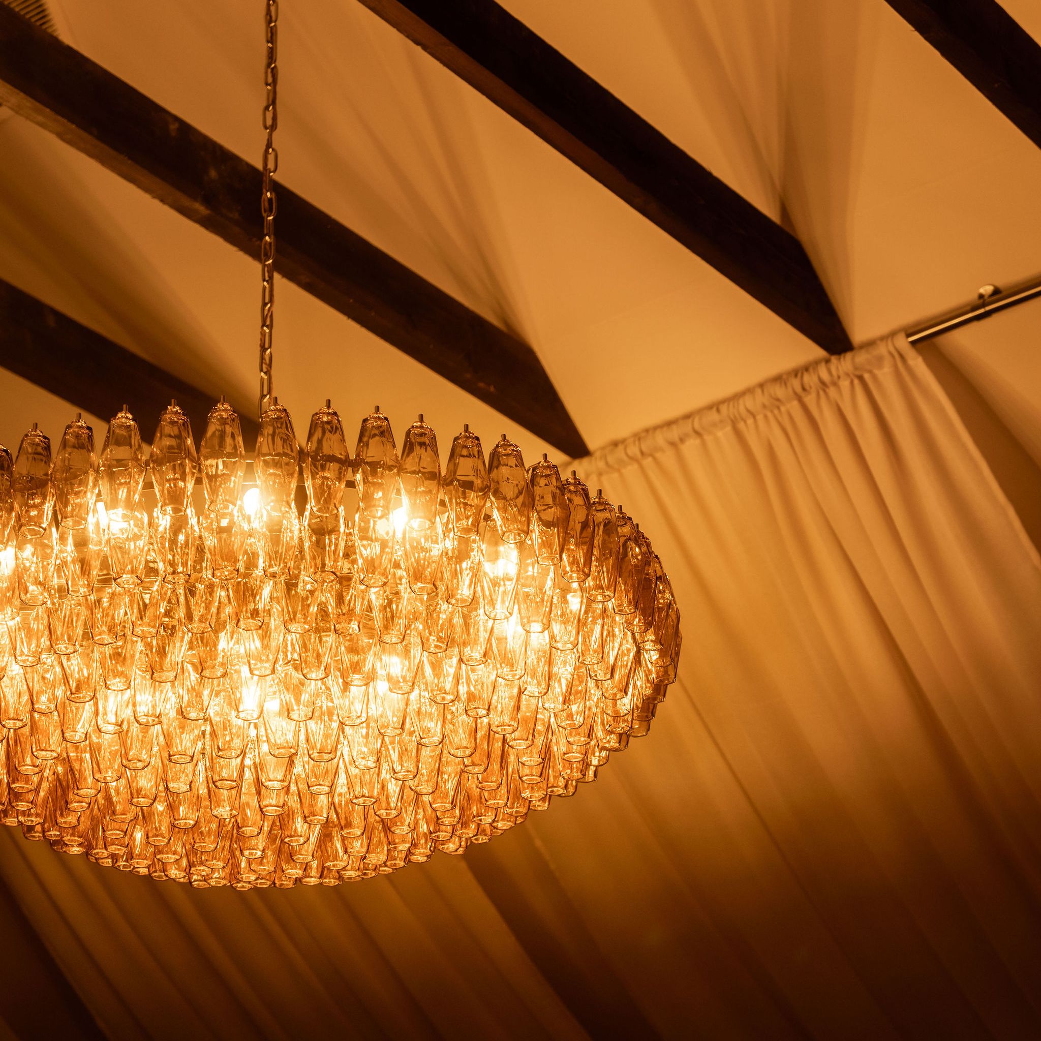 Elegant chandelier with warm yellow lights hanging from a ceiling with draped fabric and exposed beams