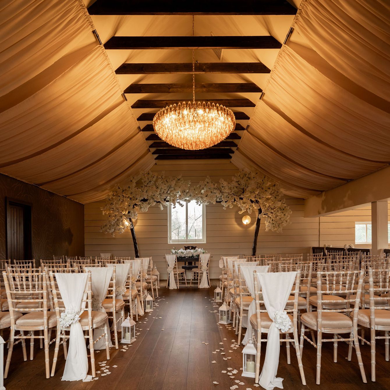 Elegant indoor wedding ceremony setup with rows of chairs, draped fabric ceiling, and a chandelier.