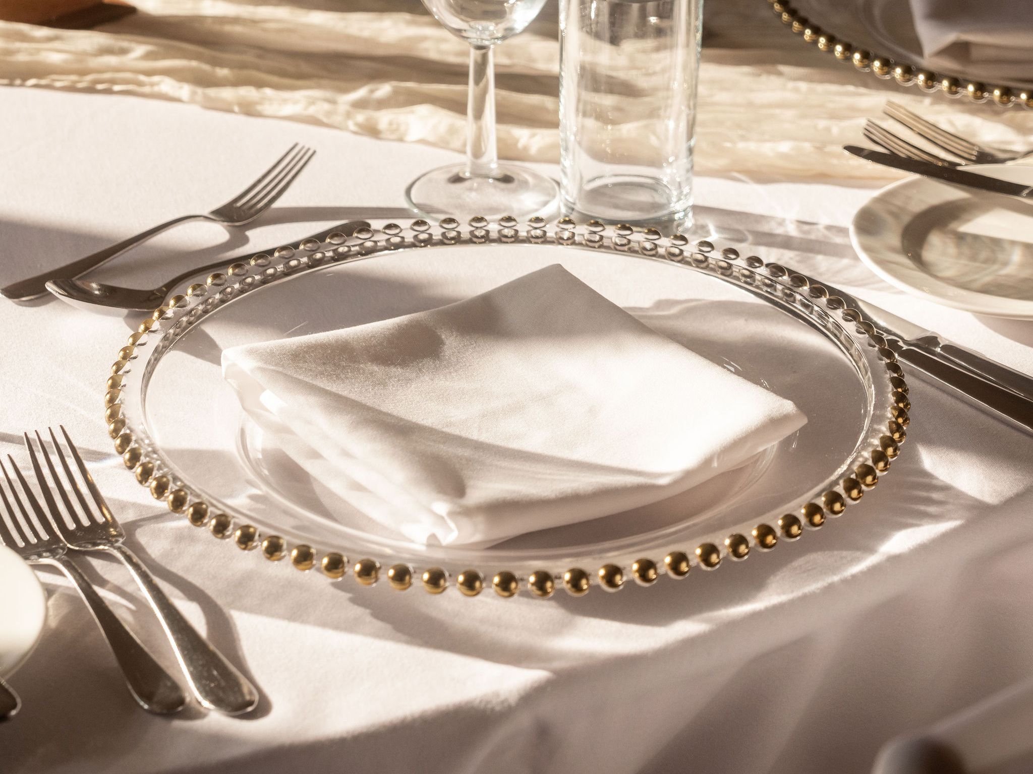 Elegant table setting with glassware, silverware, and gold-beaded charger plates on a white tablecloth.