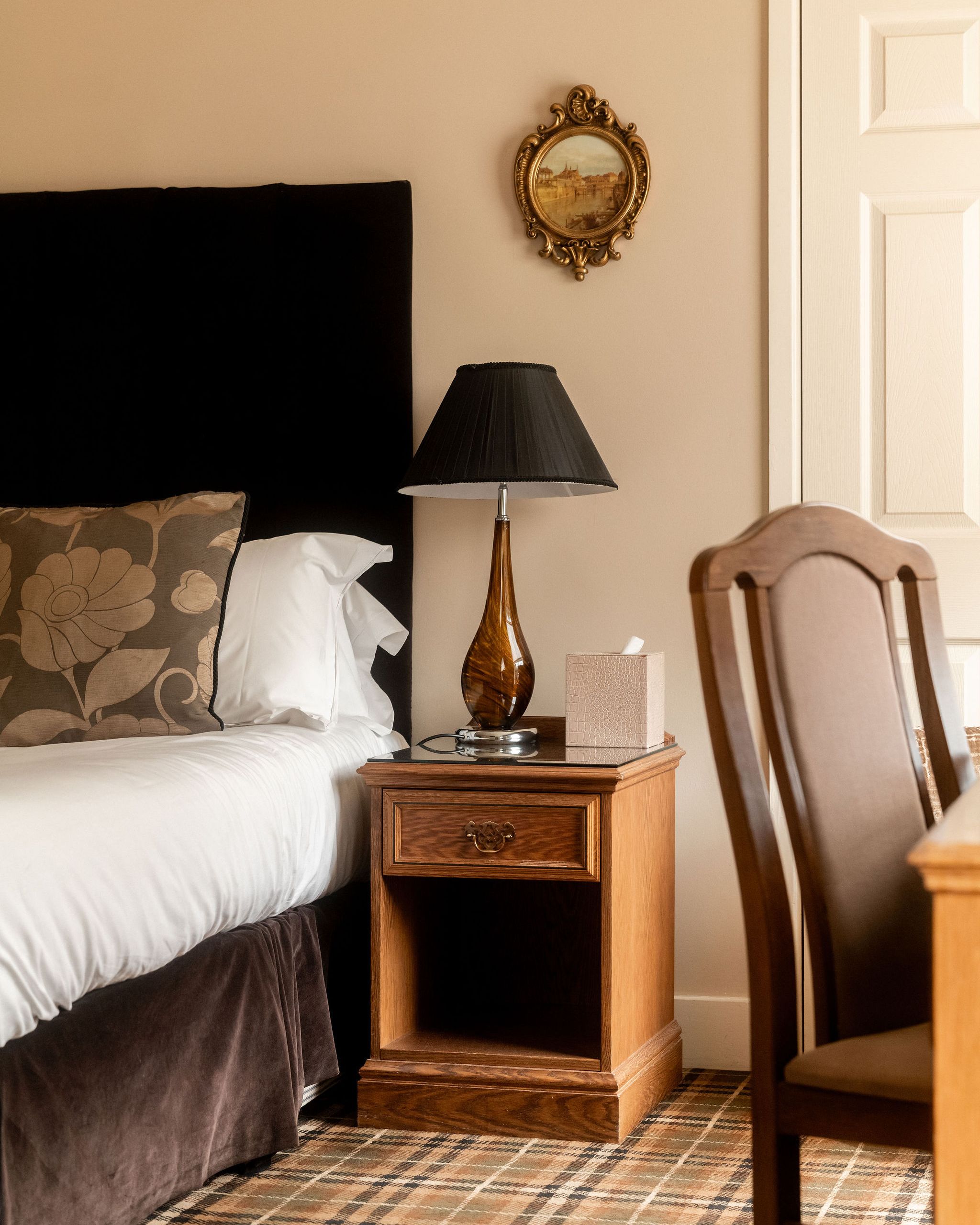 Cozy bedroom corner with a bed, floral pillow, wooden nightstand, brown table lamp, tissue box, and ornate framed painting on the wall.