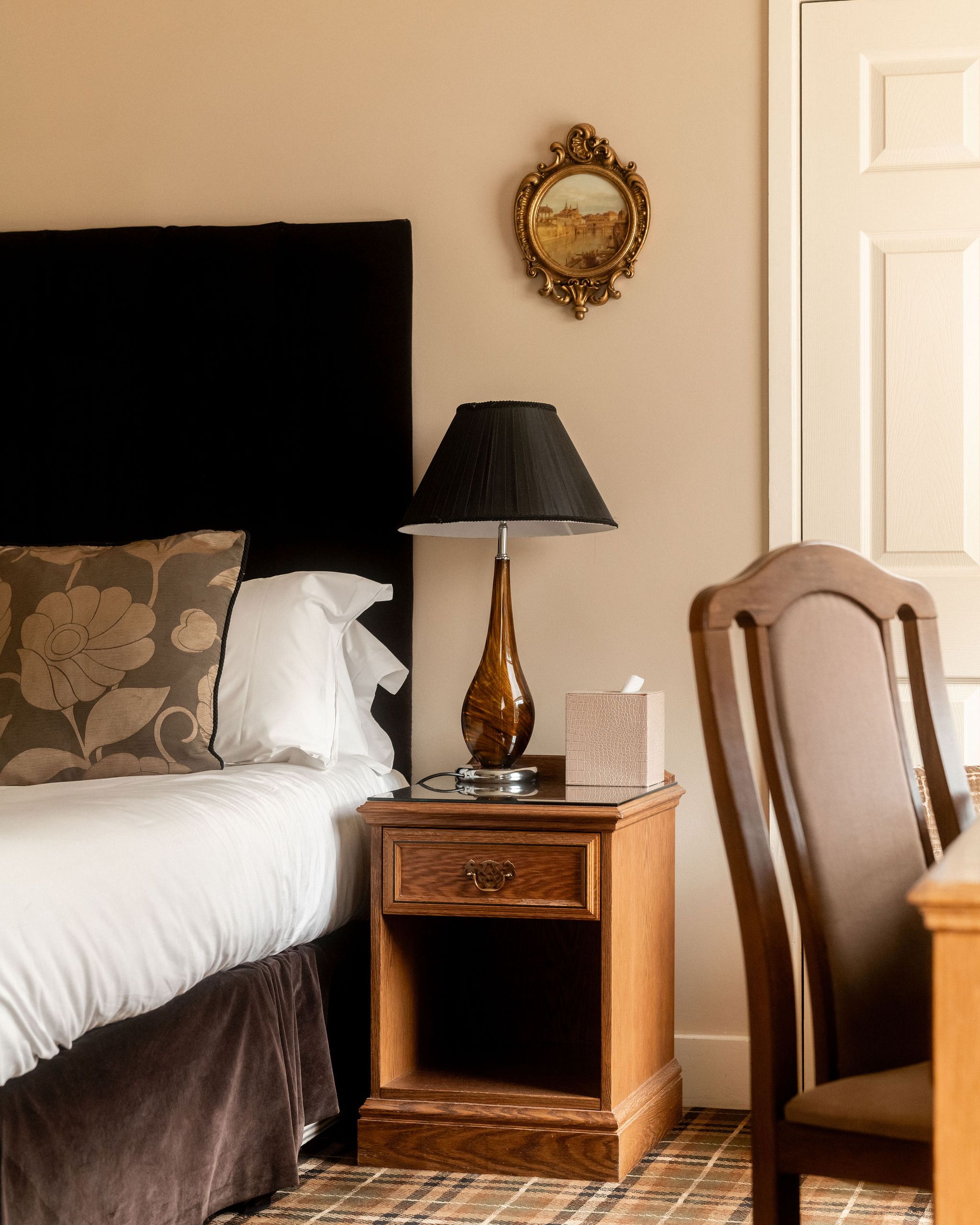 Cozy bedroom corner with a bed, floral pillow, wooden nightstand, brown table lamp, tissue box, and ornate framed painting on the wall.