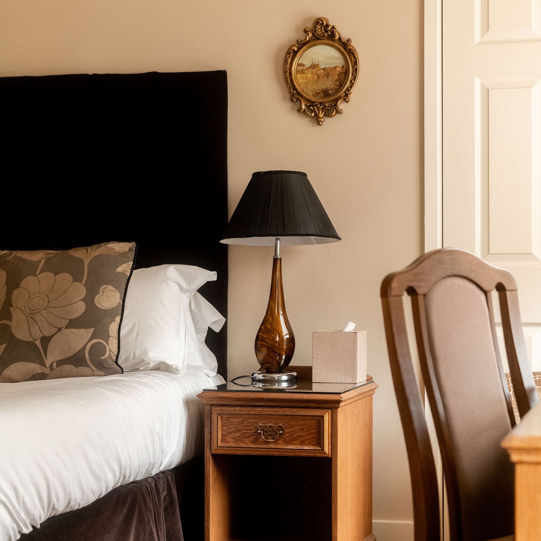 Cozy bedroom corner with a bed, floral pillow, wooden nightstand, brown table lamp, tissue box, and ornate framed painting on the wall.