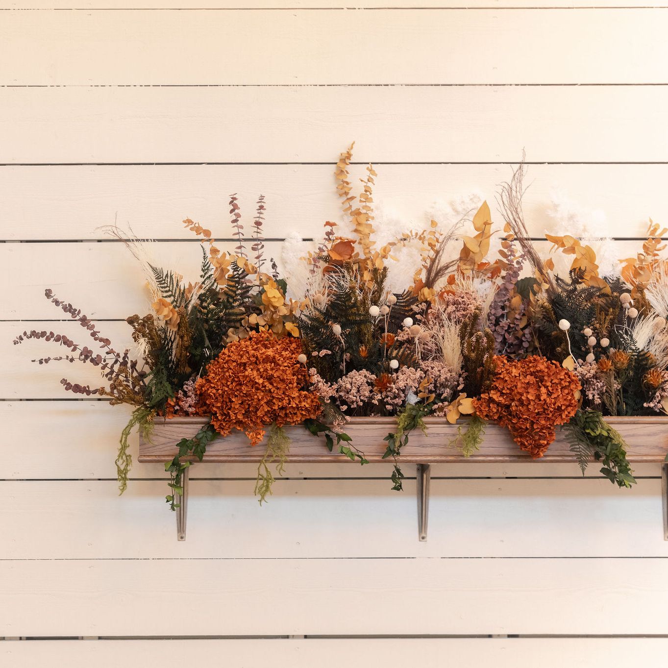Wall-mounted wooden planter box filled with dried flowers and autumnal foliage against a white shiplap wall.