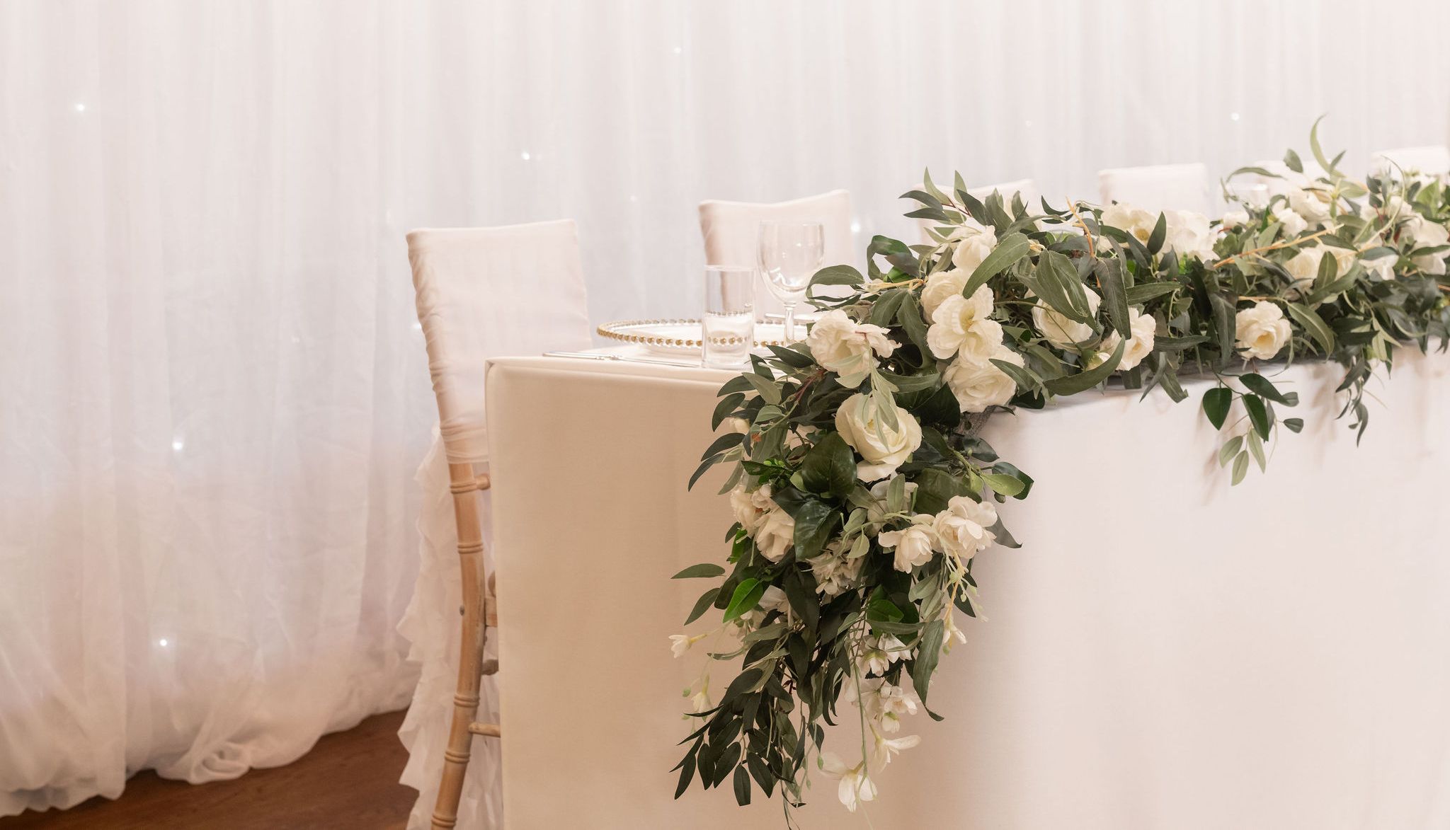Elegant wedding table with white floral centerpiece and white chairs against a sheer white backdrop
