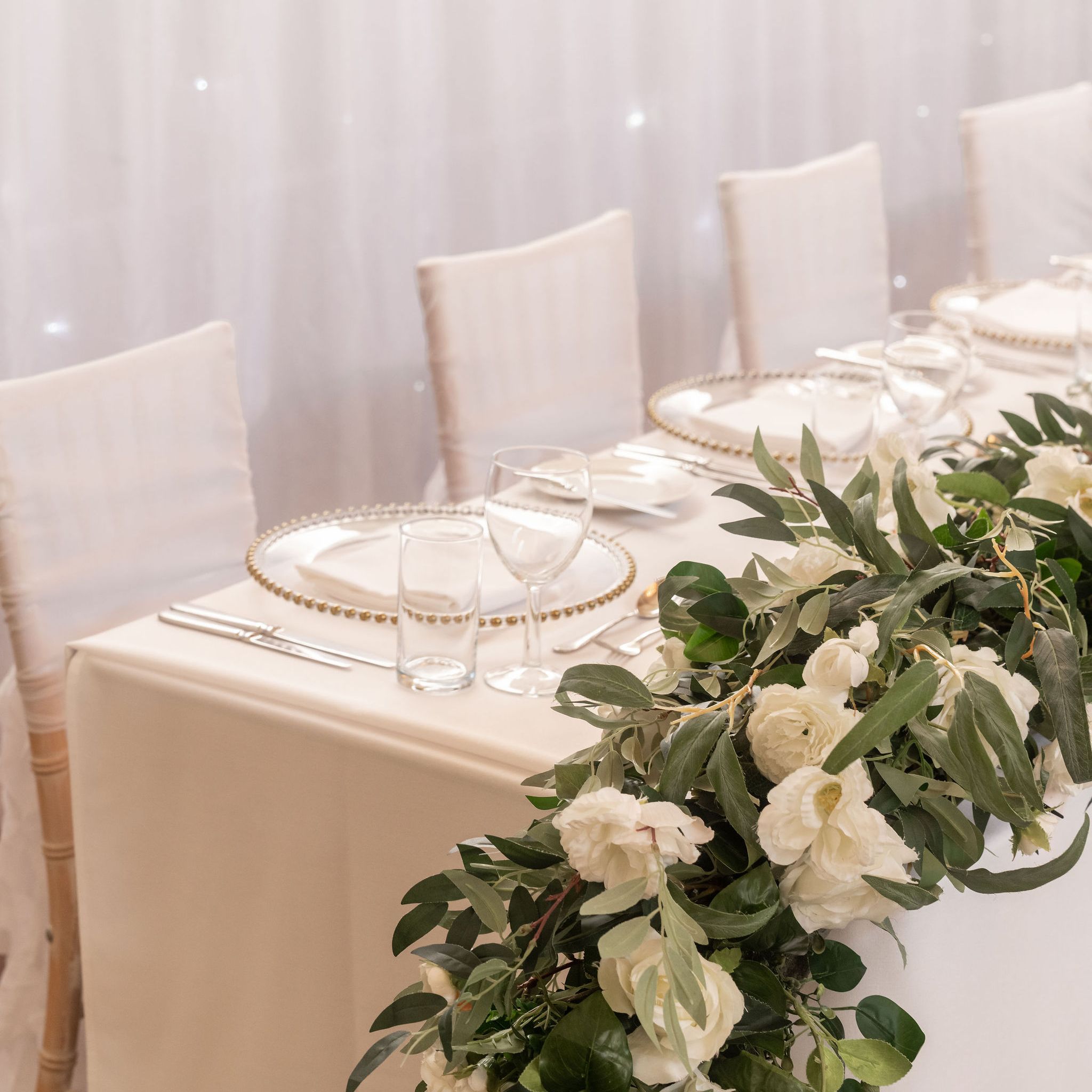 Elegant table setting with white chairs, plates, and a lush green and white floral centerpiece in front of a sheer white backdrop.