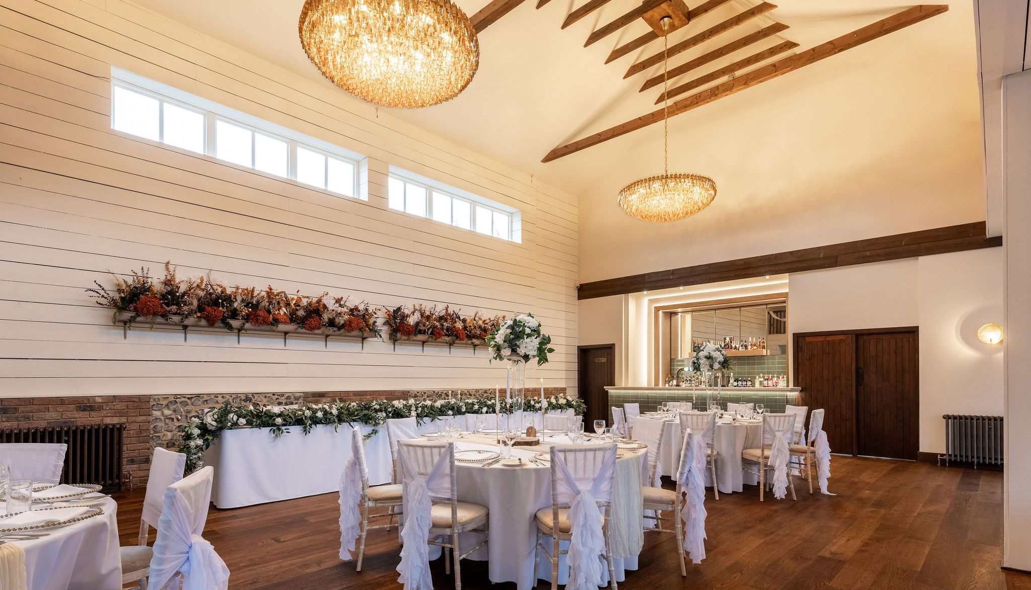Elegant dining room with round tables set for an event, decorated with white linens, floral arrangements, and chandeliers.