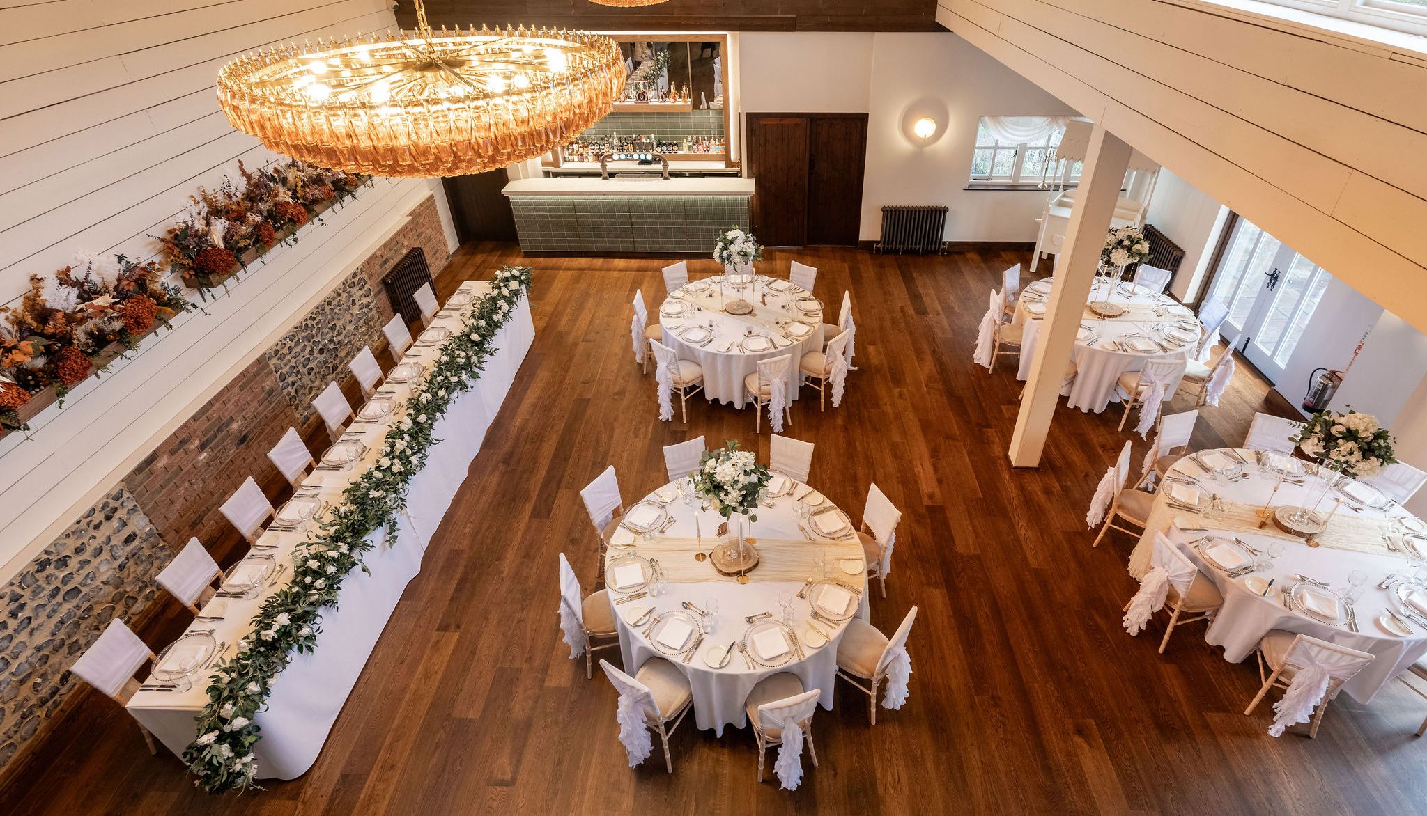 Elegant dining hall set up for an event with round tables, a long rectangular table, white chairs, and floral centerpieces.