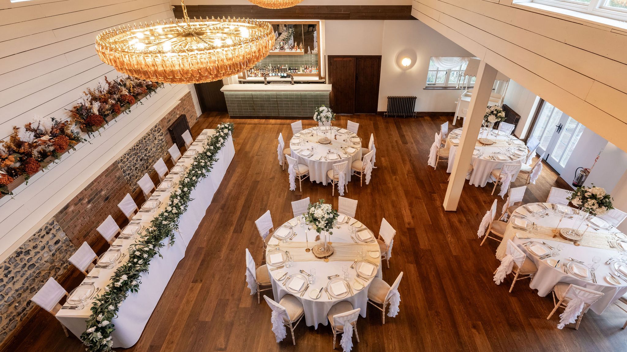 Elegant dining hall set up for an event with round tables, a long rectangular table, white chairs, and floral centerpieces.