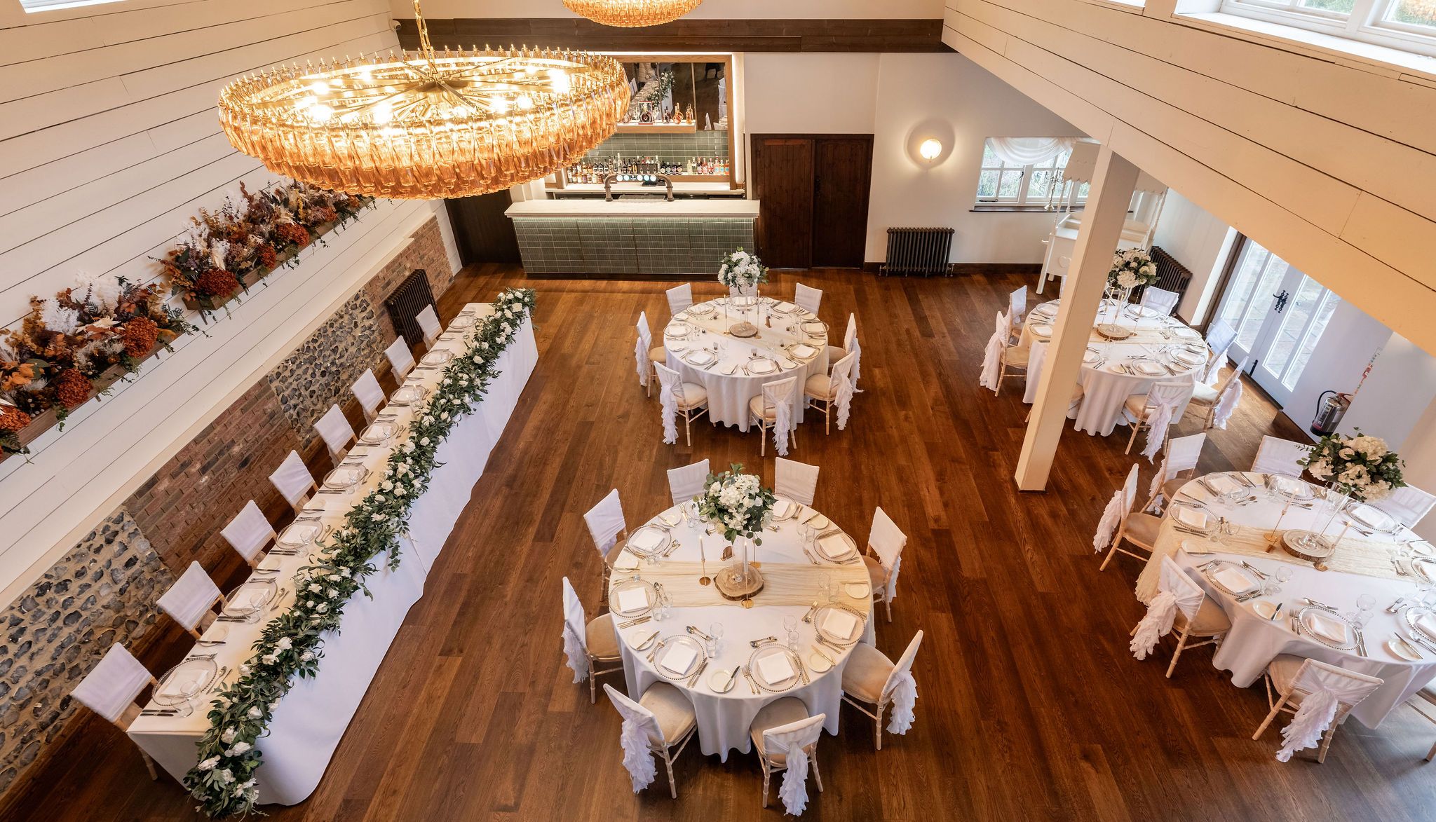 Elegant dining hall set up for an event with round tables, a long rectangular table, white chairs, and floral centerpieces.