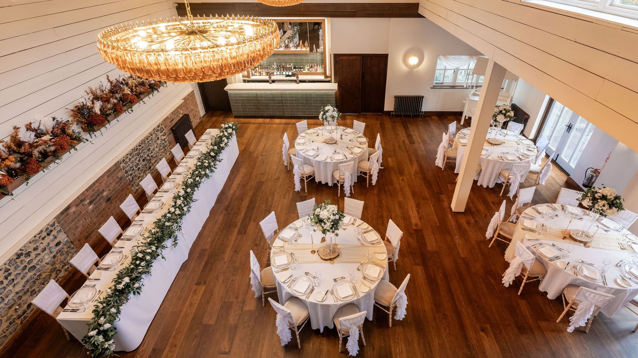 Elegant dining hall set up for an event with round tables, a long rectangular table, white chairs, and floral centerpieces.