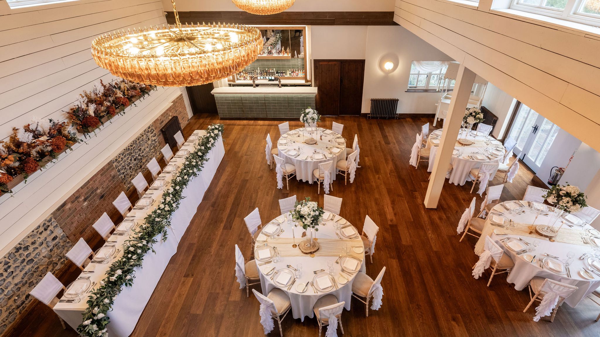 Elegant dining hall set up for an event with round tables, a long rectangular table, white chairs, and floral centerpieces.