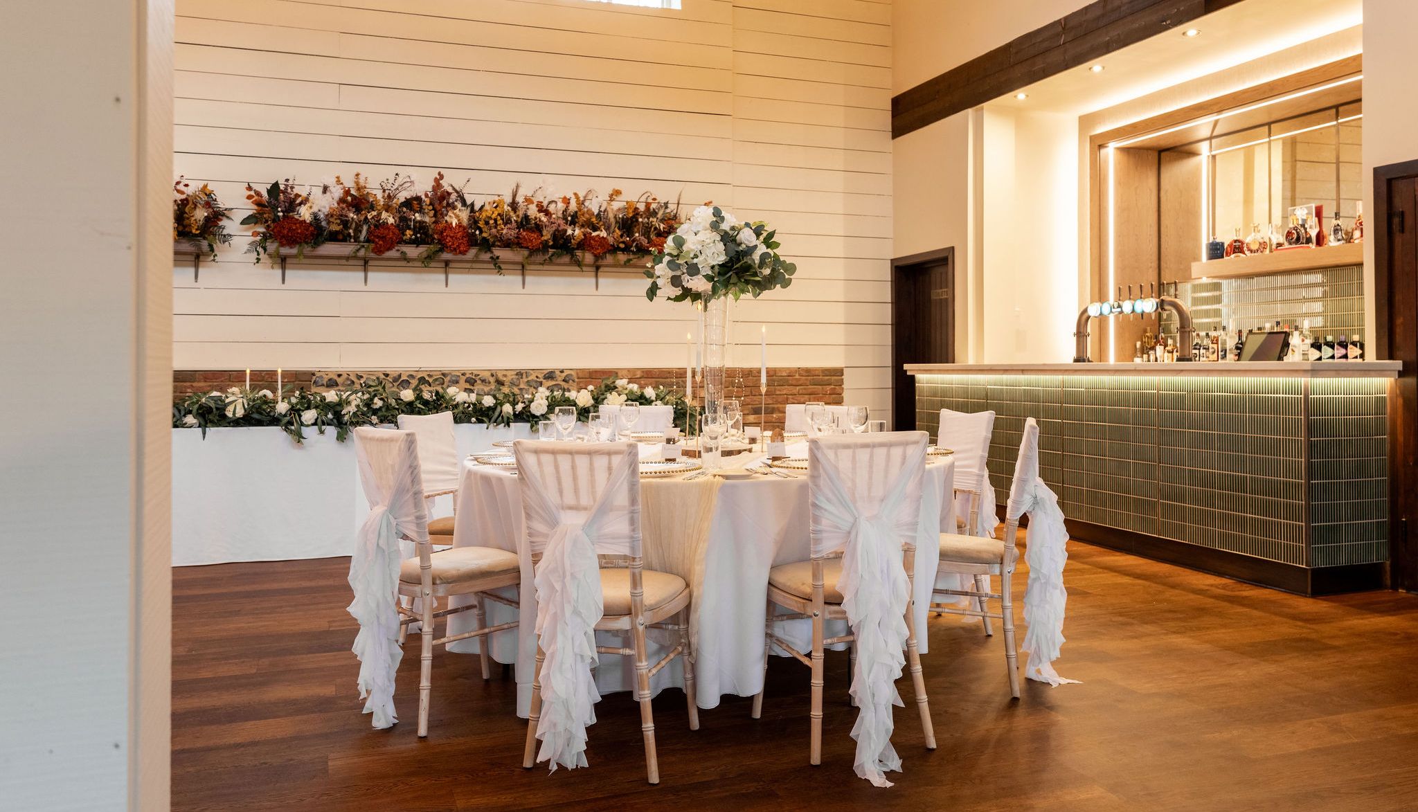 Elegant dining room with round table set for an event, featuring white chair covers and floral centerpiece, with a bar in the background.