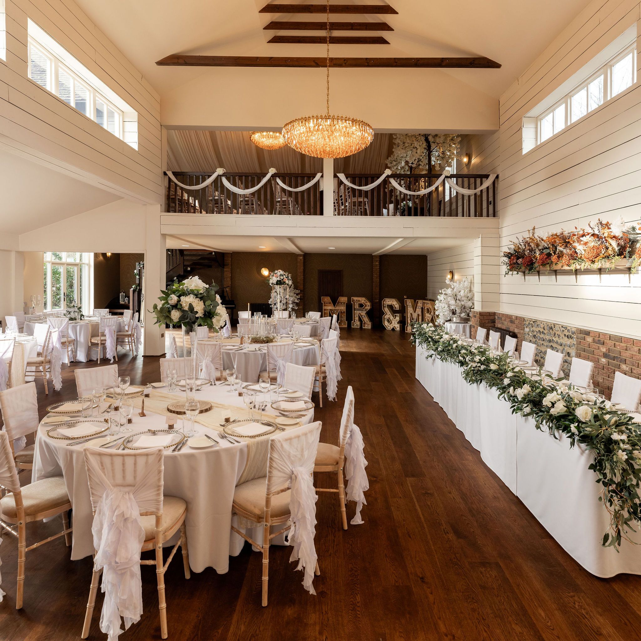 Elegant wedding reception hall with round tables, white chairs, floral centerpieces, a greenery-adorned head table, and large chandeliers.