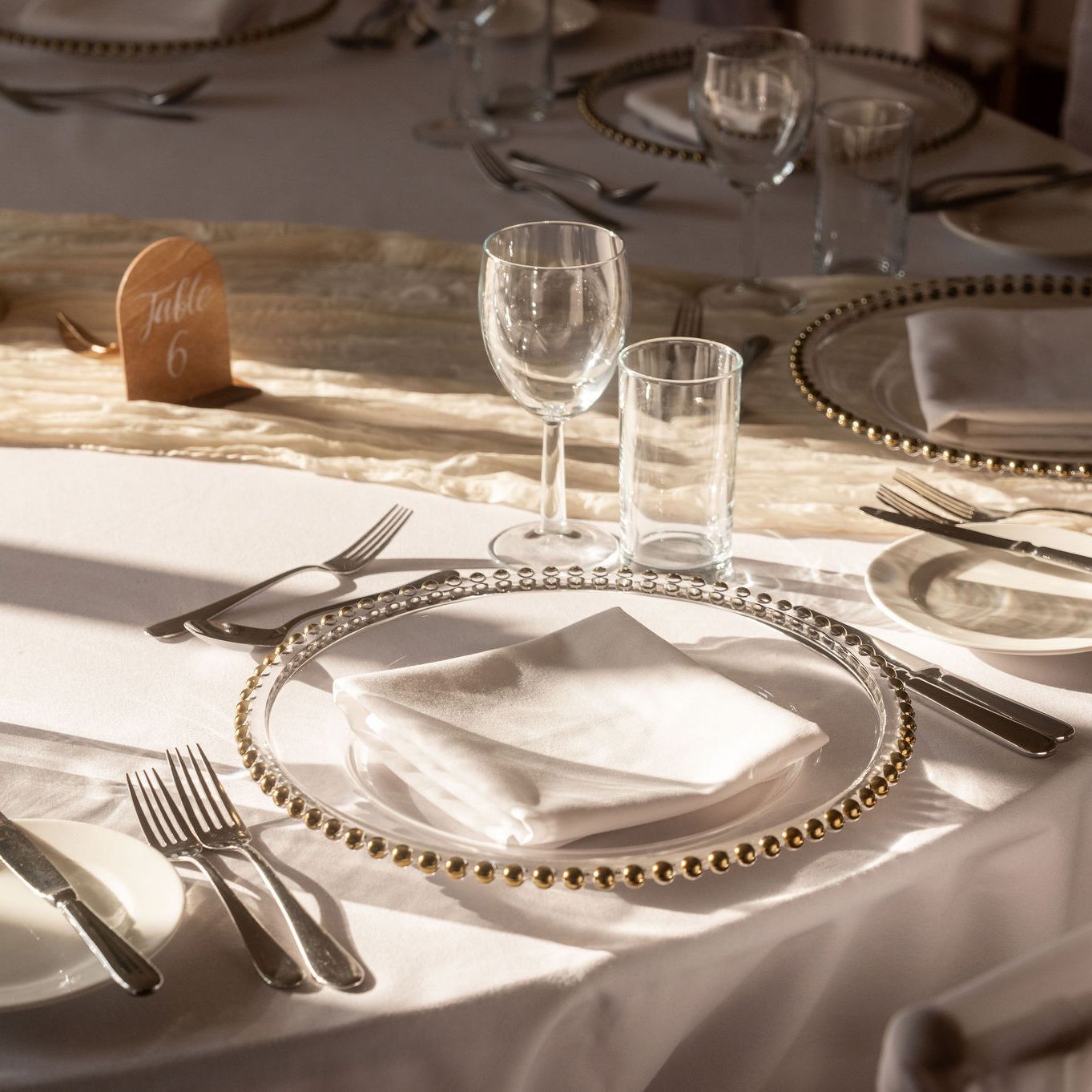 Elegant table setting with white linens, beaded charger plates, cutlery, glassware, and a table number sign.