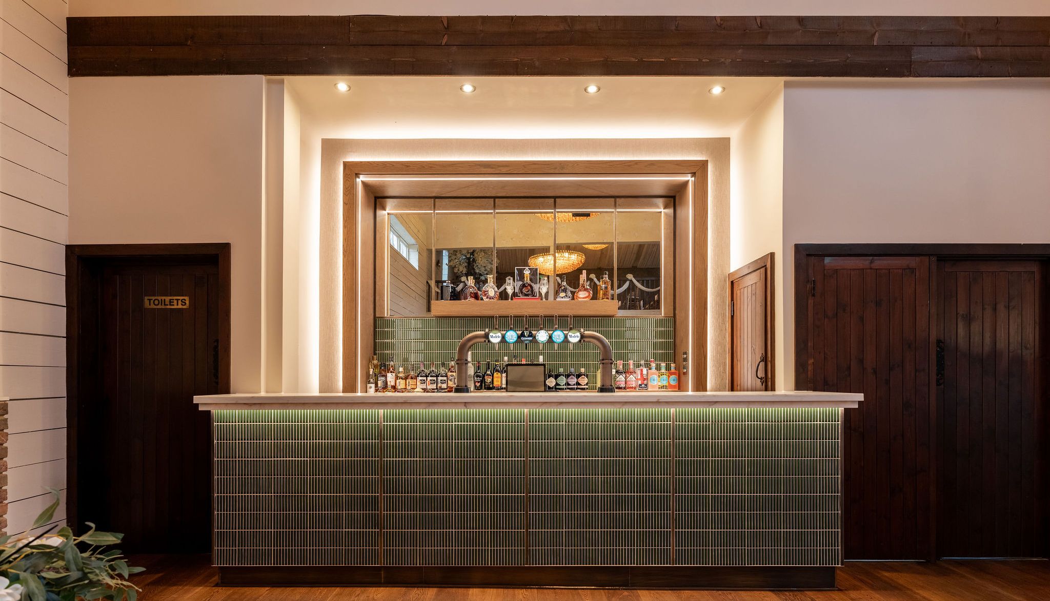 Modern bar with green tile front, bottles and beer taps, wooden accents, and a mirrored backdrop