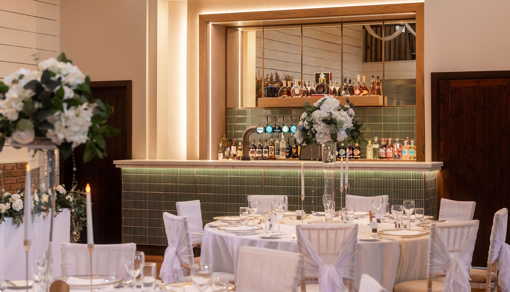 Elegant bar area with green tiles, mirrored backdrop, and wedding reception tables in front with white floral centerpieces.