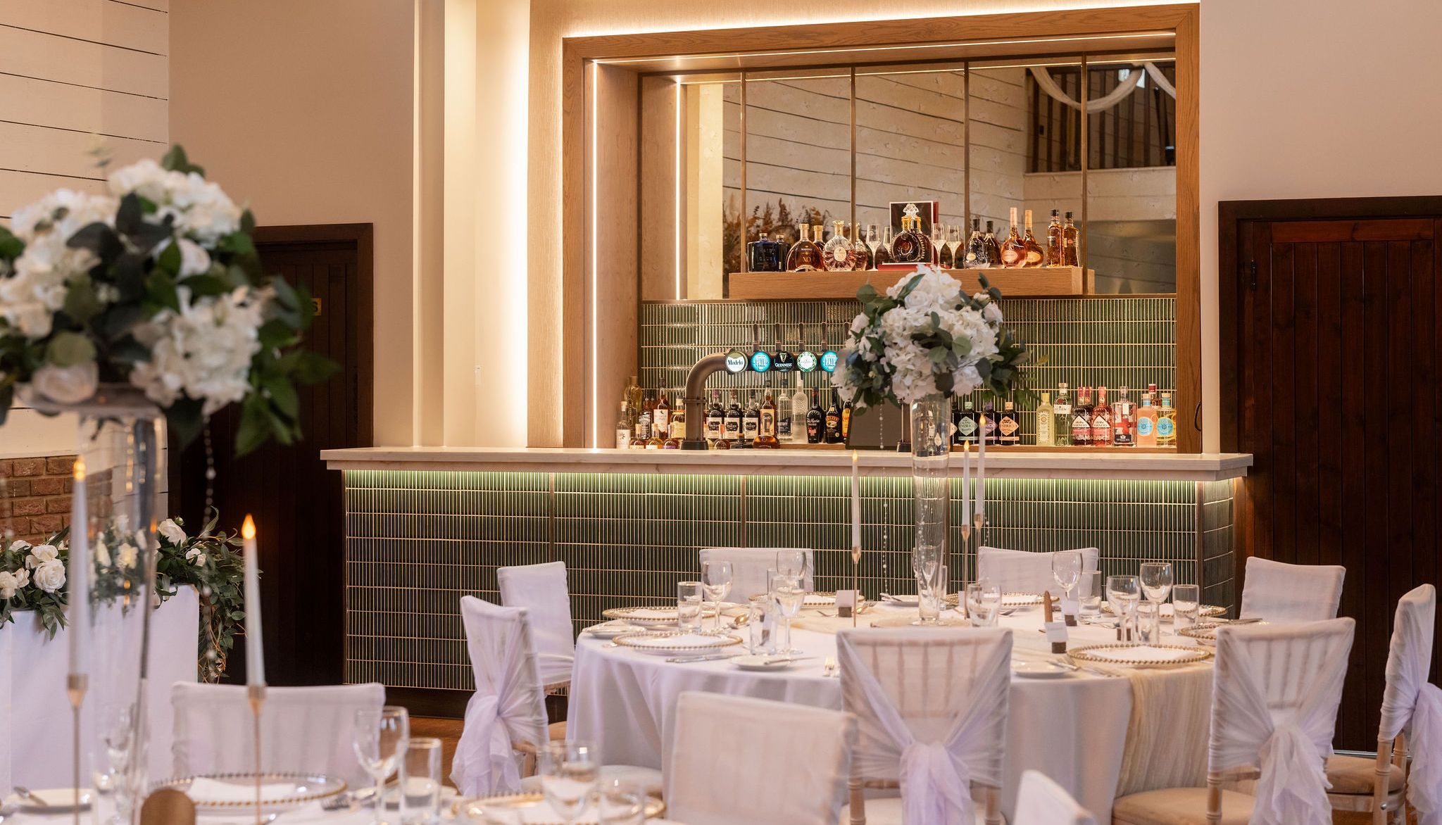 Elegant bar area with green tiles, mirrored backdrop, and wedding reception tables in front with white floral centerpieces.