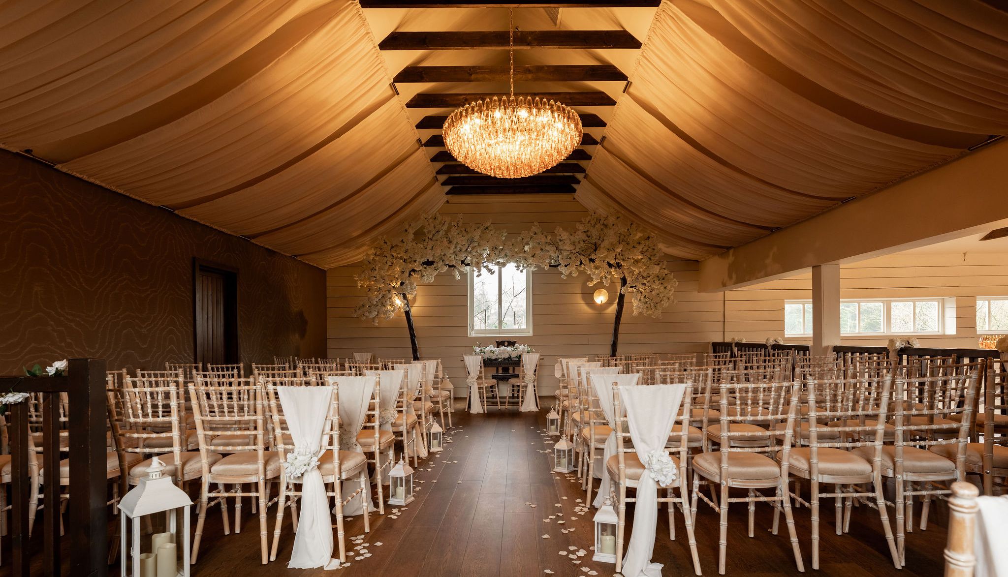 Elegant indoor wedding ceremony setup with draped ceiling, chandelier, and rows of chairs decorated with white fabric and flowers.