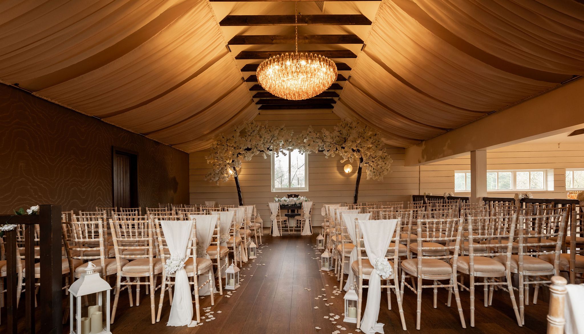 Elegant indoor wedding ceremony setup with draped ceiling, chandelier, and rows of chairs decorated with white fabric and flowers.