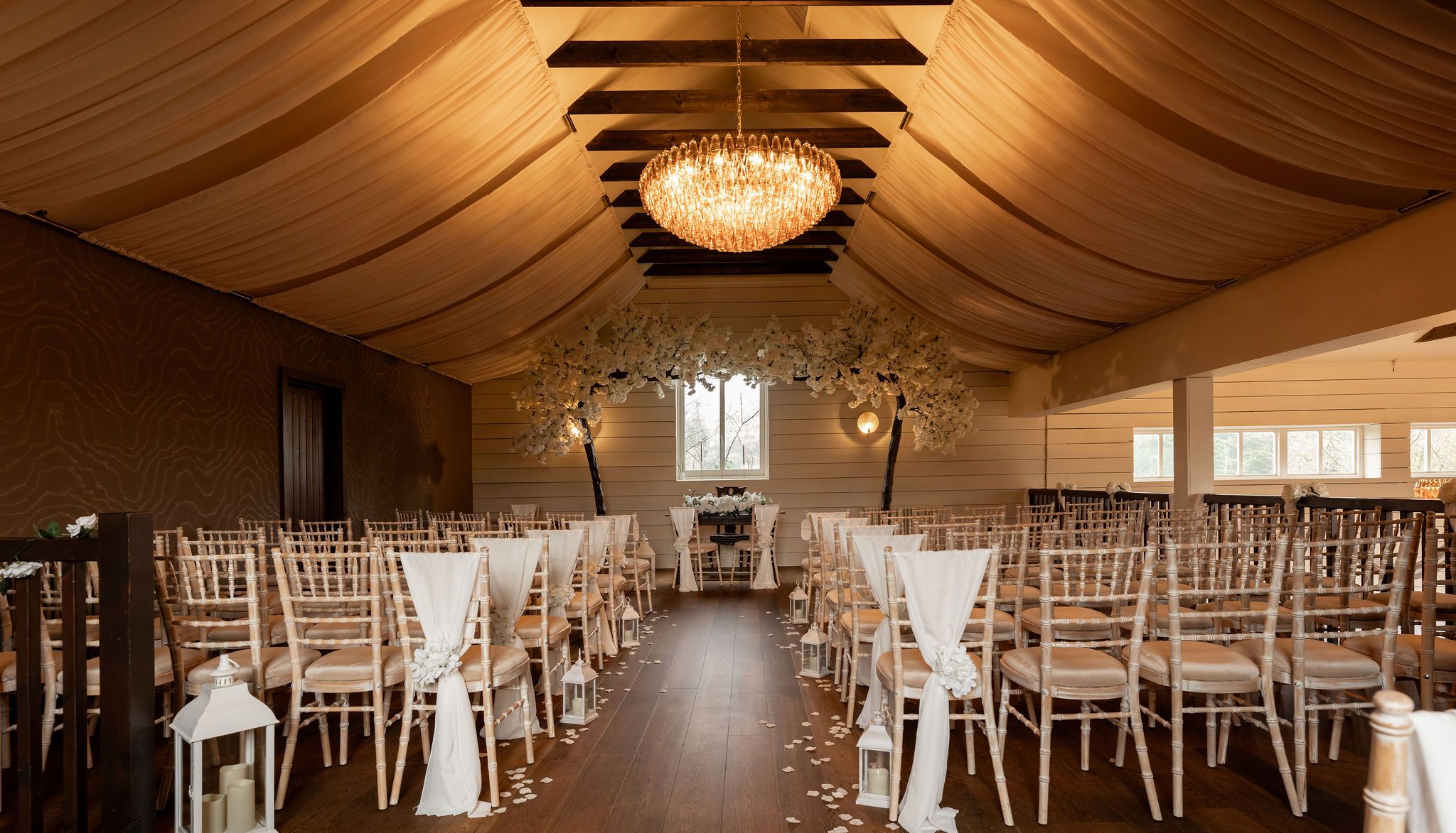 Elegant indoor wedding ceremony setup with draped ceiling, chandelier, and rows of chairs decorated with white fabric and flowers.