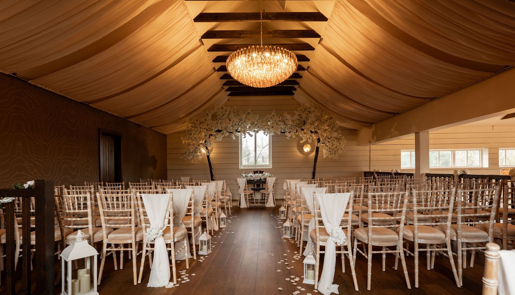 Elegant indoor wedding ceremony setup with draped ceiling, chandelier, and rows of chairs decorated with white fabric and flowers.