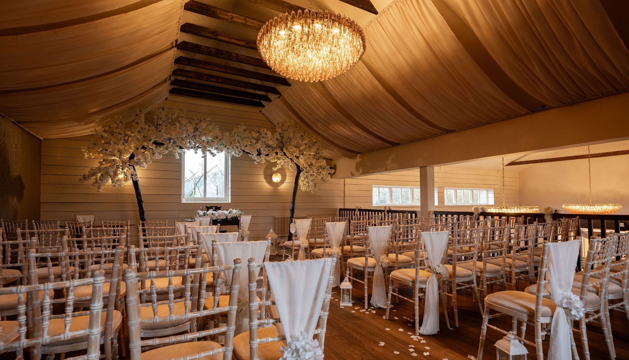 Elegant indoor wedding ceremony setup with rows of chairs, white drapes, flower arrangements, and chandeliers.