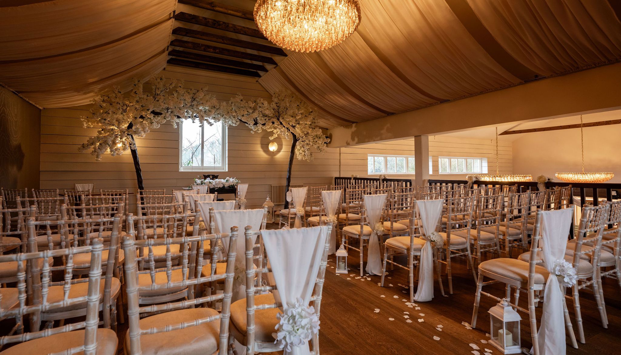 Elegant indoor wedding ceremony setup with rows of chairs, white drapes, flower arrangements, and chandeliers.