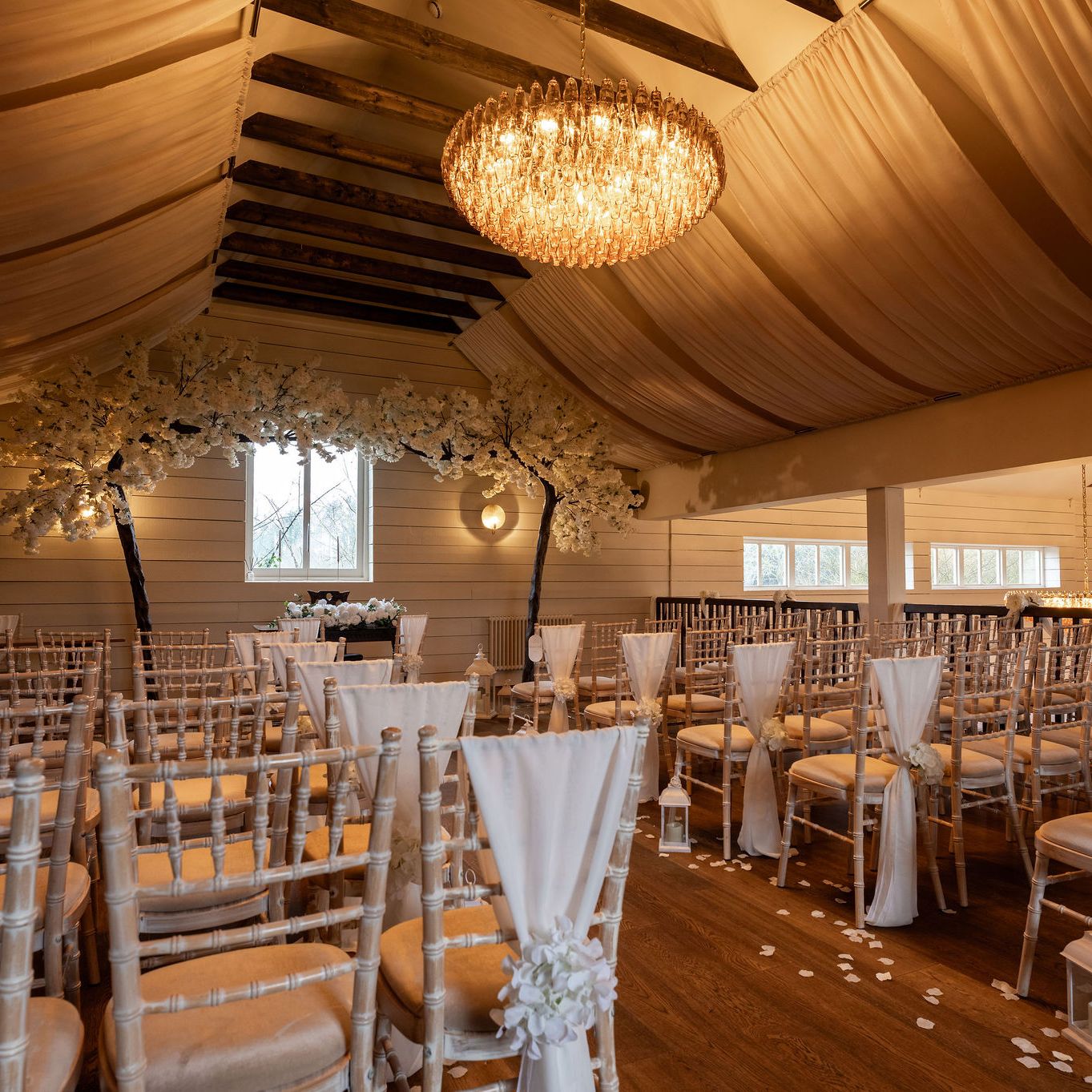Elegant indoor wedding ceremony setup with rows of chairs, white drapes, flower arrangements, and chandeliers.
