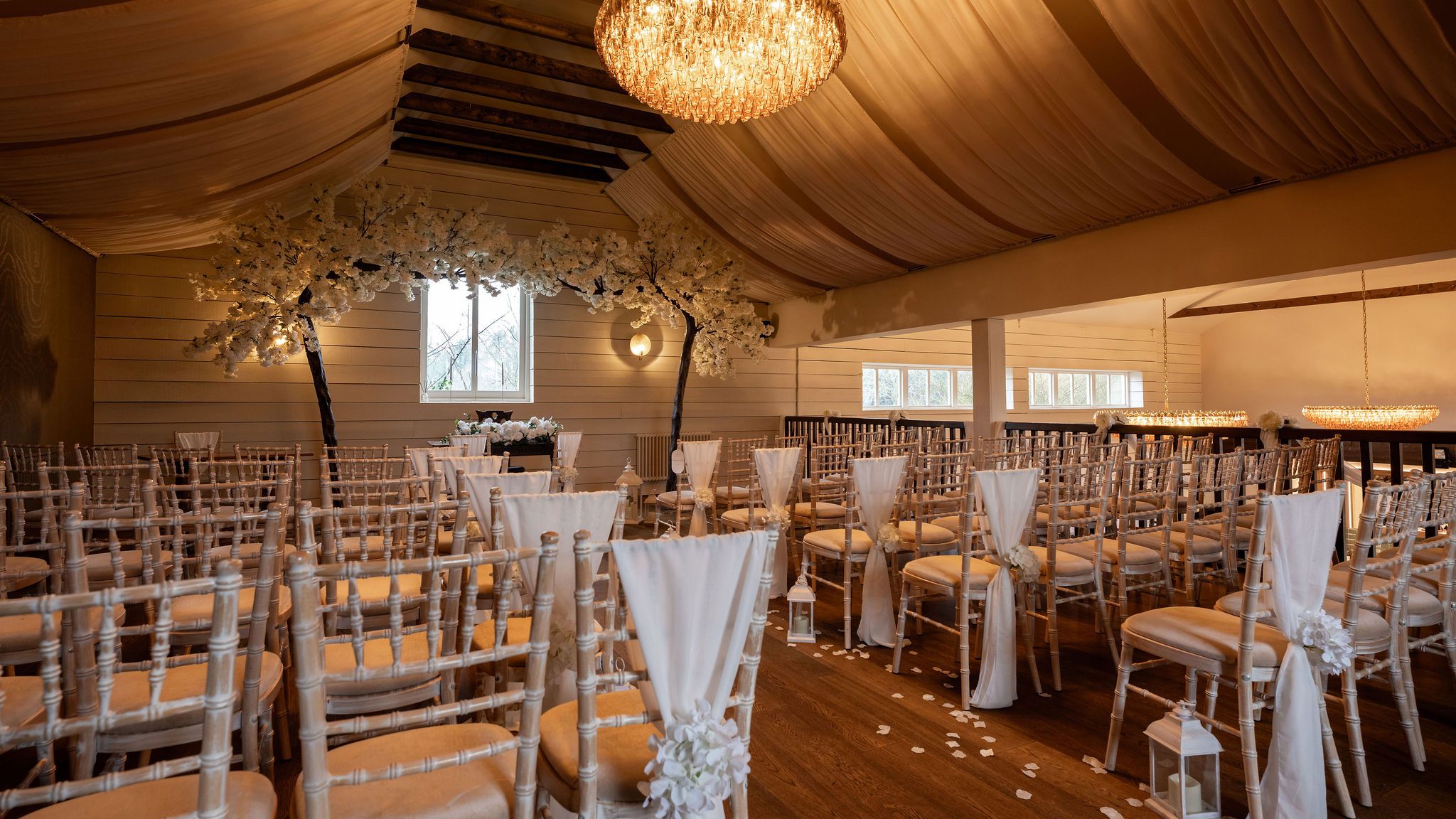 Elegant indoor wedding ceremony setup with rows of chairs, white drapes, flower arrangements, and chandeliers.