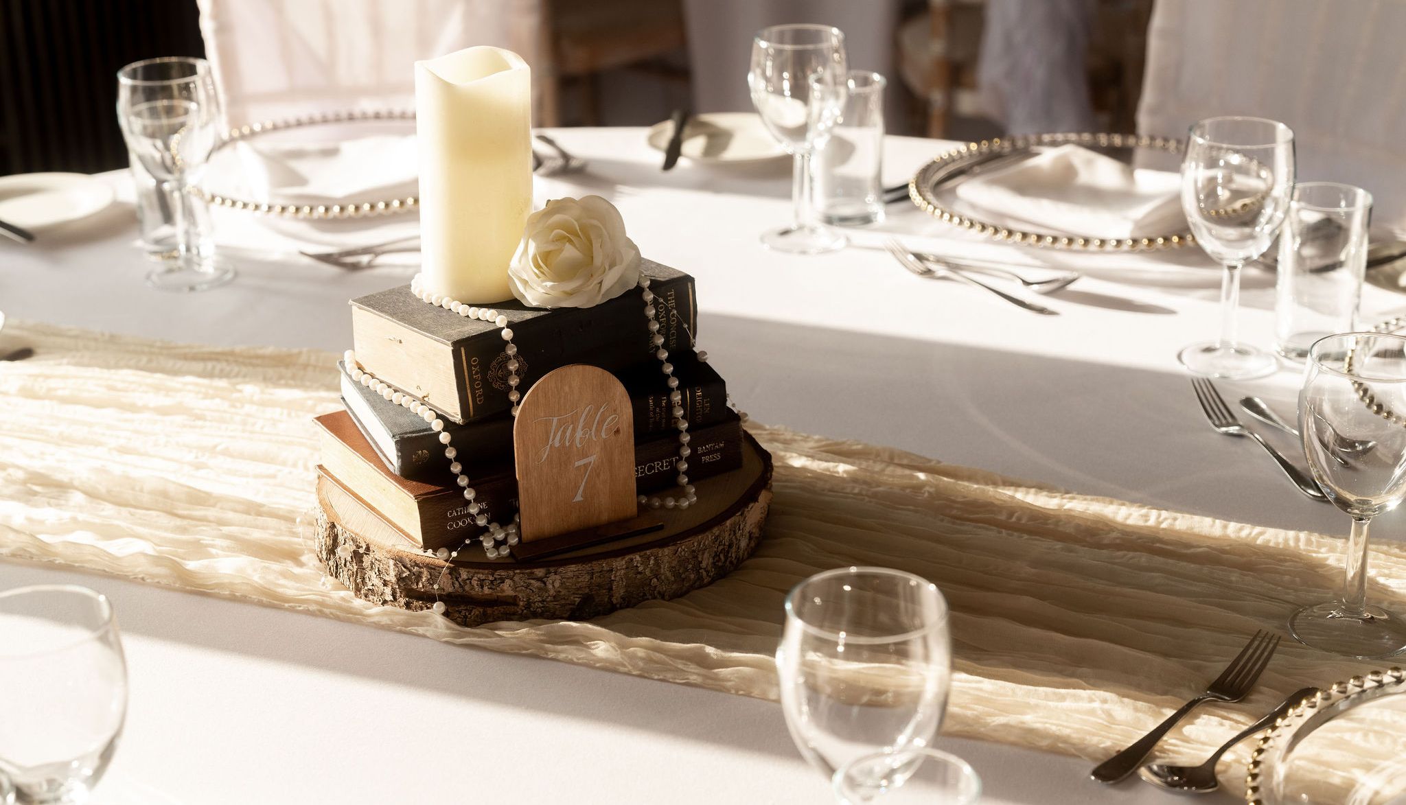 Elegant table centerpiece with stacked books, a candle, a white rose, pearl beads, and a wooden table number surrounded by tableware.