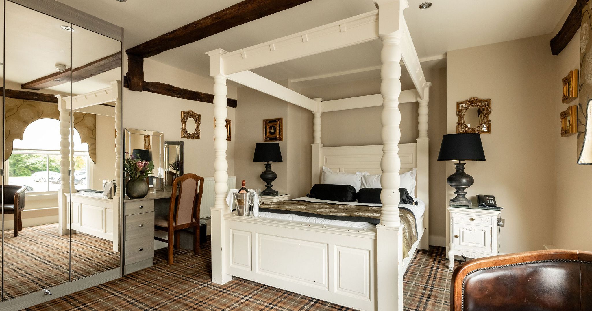 A well-decorated hotel room featuring a white four-poster bed, black lamps, a vanity with mirror and chair, plaid carpet, and classic framed mirrors on cream-colored walls.