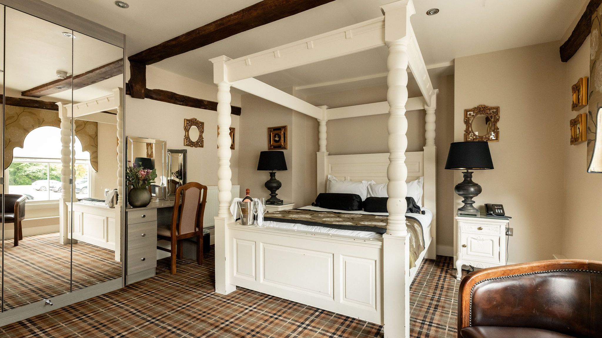 A well-decorated hotel room featuring a white four-poster bed, black lamps, a vanity with mirror and chair, plaid carpet, and classic framed mirrors on cream-colored walls.