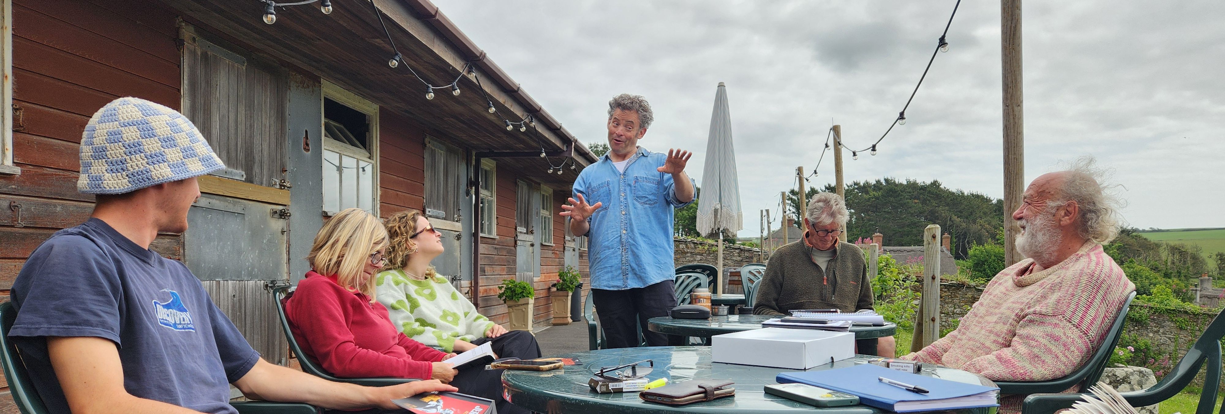 A group of people sitting outside around a table, engaged in a lively discussion or meeting, with one person standing and gesturing as they speak.