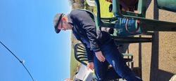 Man in a flat cap and glasses sitting on a green plastic chair outdoors, reading papers, with a blue sky and countryside in the background.