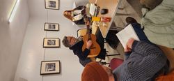 A man playing guitar in a small room with people reading books around a table.