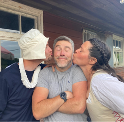 Three adults pose in front of a rustic wooden building, with a man in the middle being kissed on each cheek by a woman and a man in costume.