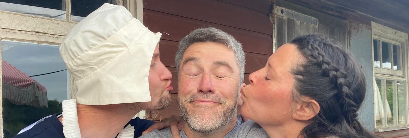 Three adults pose in front of a rustic wooden building, with a man in the middle being kissed on each cheek by a woman and a man in costume.